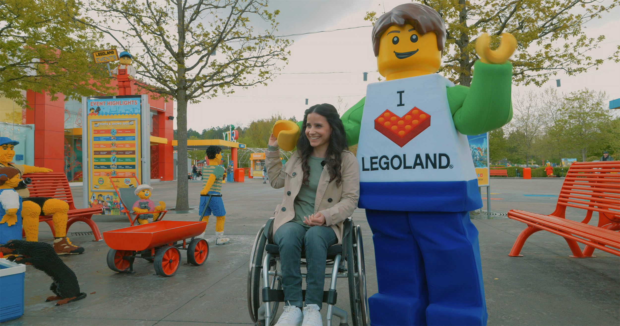 Junge Frau im Rollstuhl und ein LEGO Charakter im Eingangsbereich des LEGOLAND Deutschlands, wobei die Frau lacht, den LEGO Charakter an der Hand hält und dieser seinen Arm um ihre Schulter gelegt hat und mit dem anderen Arm nach vorne winkt.