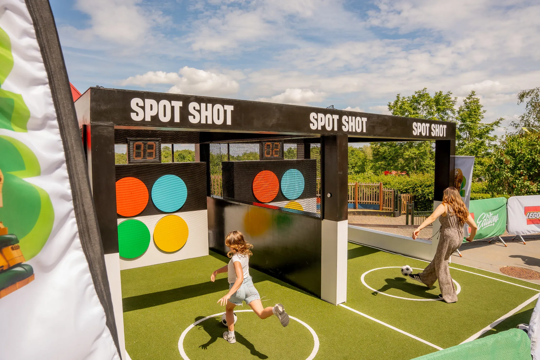 Zwei Personen spielen auf einem Spielfeld mit Torwand und bunten Zielscheiben unter dem Schriftzug »SPOT SHOT«