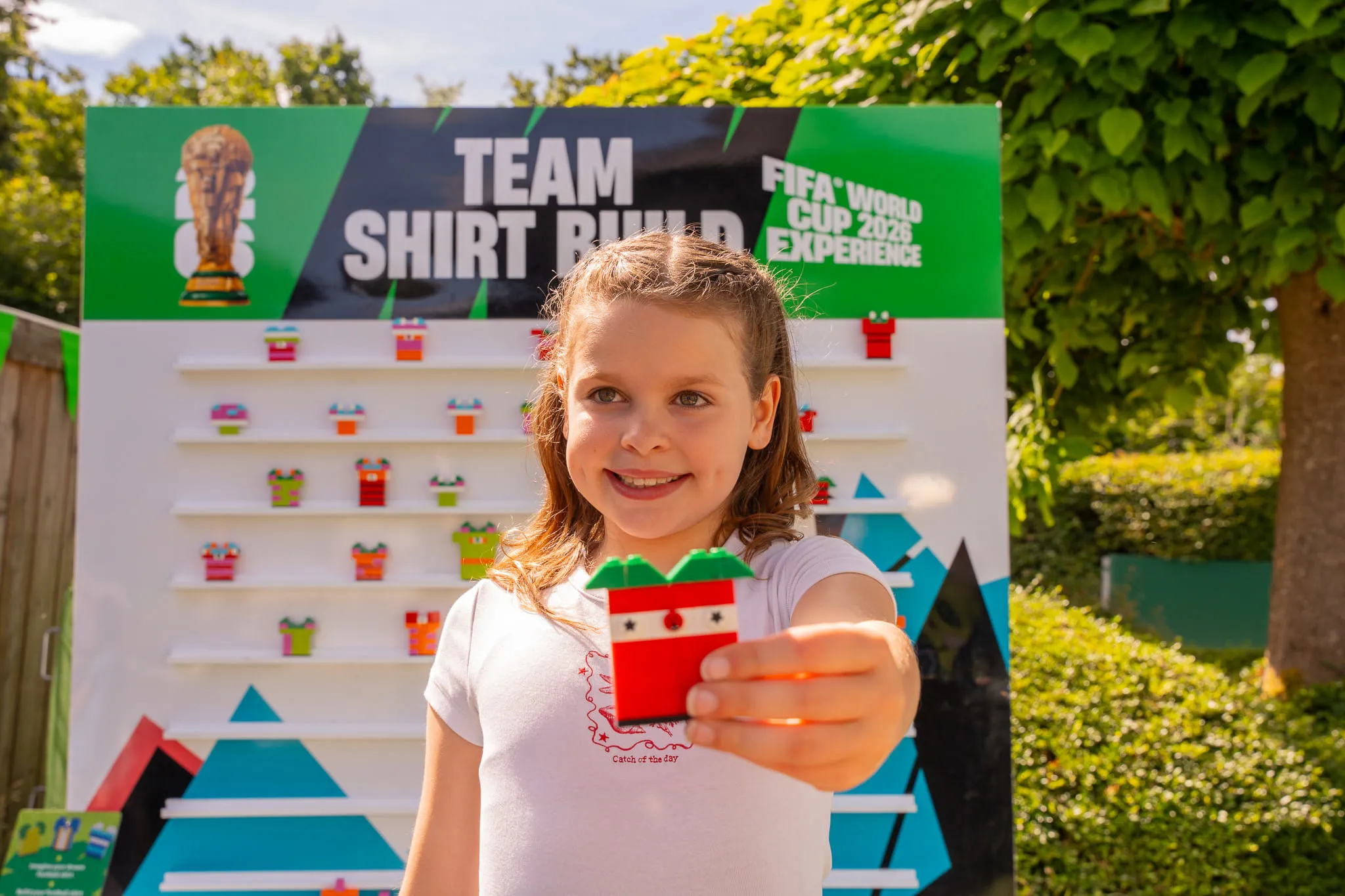 Kind hält ein rotes Trikot aus LEGO Steinen vor einem Spielstand mit der Aufschrift 'TEAM SHIRT BUILD FIFA WORLD CUP 2026 EXPERIENCE'