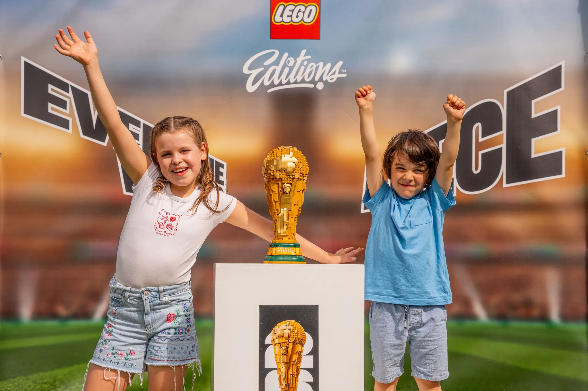 Zwei Kinder mit erhobenen Armen stehen neben einer LEGO-Nachbildung des FIFA-Weltpokals auf einem Podest vor einem Banner mit dem LEGO-Logo und dem Schriftzug 'Editions'