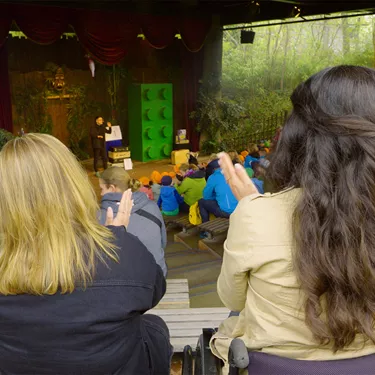Zwei Frauen, die im Affentheater des LEGOLAND Deutschlands klatschen, wobei die Frau links auf einer Bank sitzt und die andere Frau rechts in einem Rollstuhl sitzt, während im Hintergrund eine Bühne, auf der ein Mann steht, und das weitere Publikum zu sehen ist.