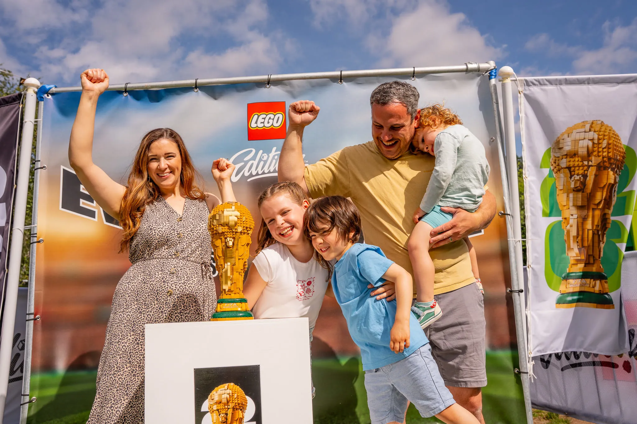 Familie mit zwei Kindern jubelt vor einer LEGO Weltmeisterschaftstrophäe aus LEGO Steinen auf einer Veranstaltung im Freien