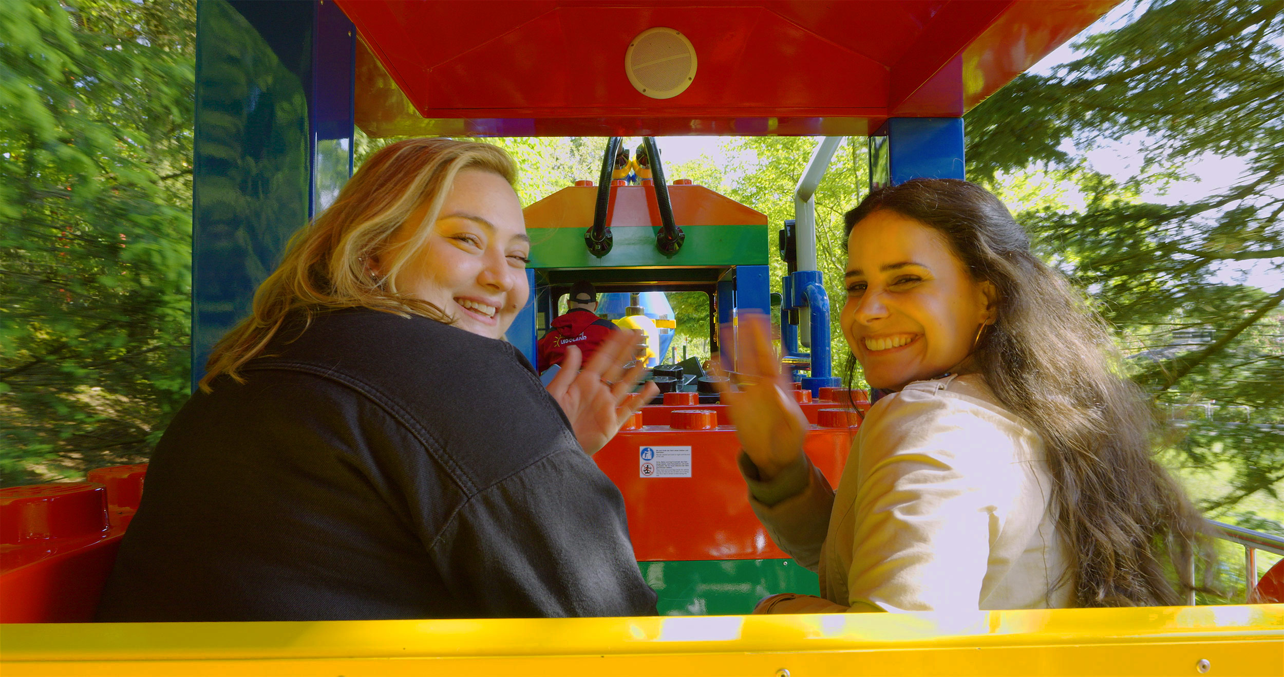 Zwei lachende Frauen, die eine Parkrundfahrt im LEGOLAND Express, einem bunten Zug, von LEGOLAND Deutschland machen und sich nach hinten drehen, um in die Kamera zu winken.