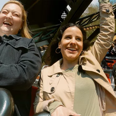 Zwei Frauen, die in der Achterbahn "Drachenjagd" des LEGOLAND Deutschlands sitzen und lachen, wobei die Frau rechts ihre rechte Hand festhält und ihre linke Hand in die Luft hebt, während im Hintergrund die Wagen des Fahrgeschäfts und die Schienen der Achterbahn zu sehen sind.