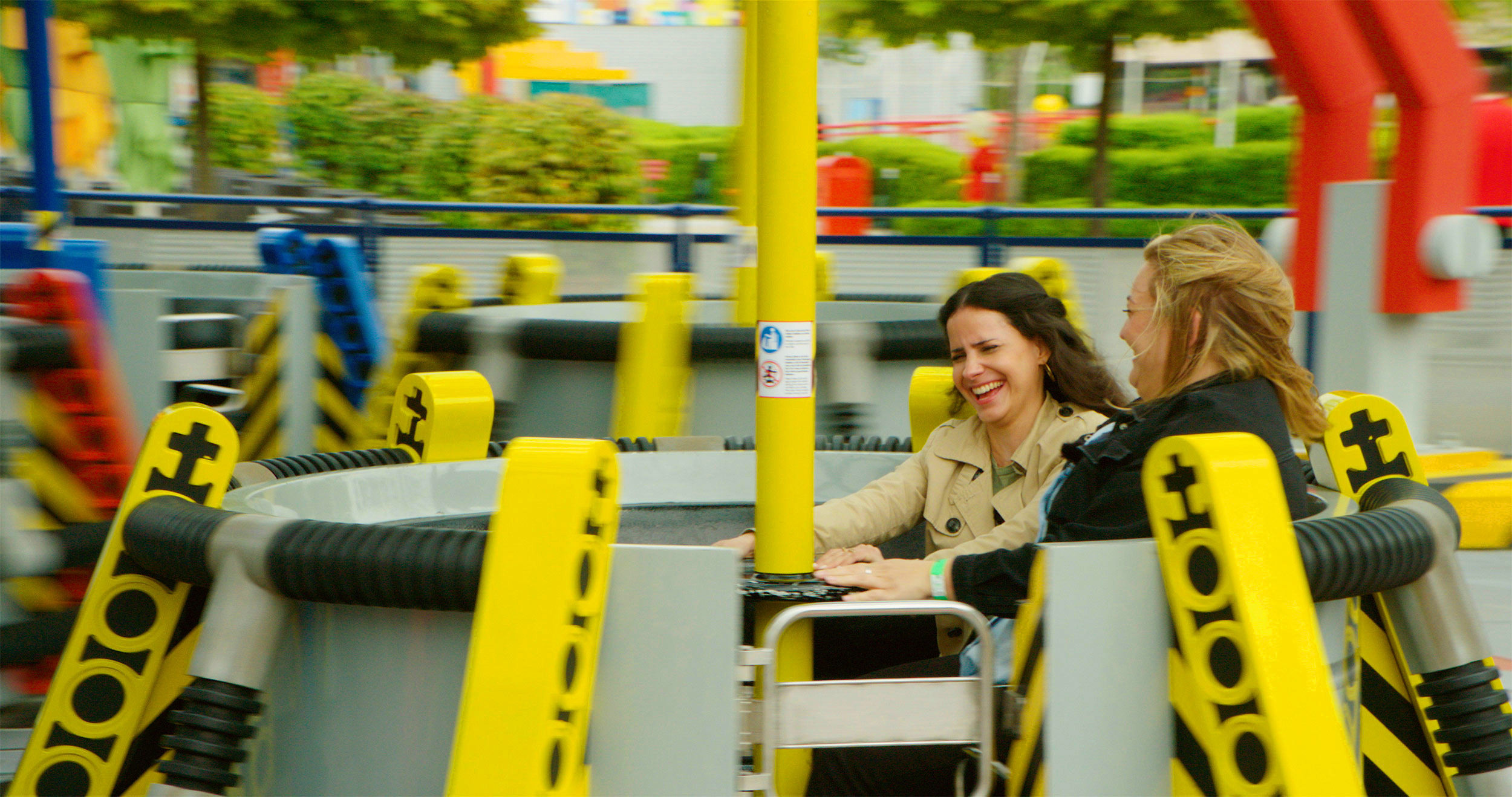 Zwei Frauen, die in der Technoschleuder des LEGOLAND Deutschlands sitzen, während sie ihre Hände an der Stange in der Mitte festhalten und lachen. Das Fahrgeschäft besteht aus mehreren runden Elementen auf Drehscheiben mit einer Stange in der Mitte.