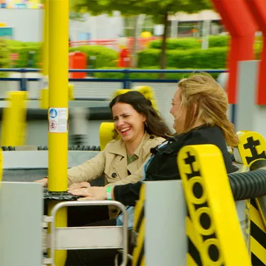 Zwei Frauen, die in der Technoschleuder des LEGOLAND Deutschlands sitzen, während sie ihre Hände an der Stange in der Mitte festhalten und lachen. Das Fahrgeschäft besteht aus mehreren runden Elementen auf Drehscheiben mit einer Stange in der Mitte.