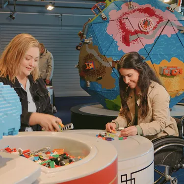 Zwei Frauen - wobei einer der Frauen im Rollstuhl sitzt - die im "Rebuild the World" von LEGOLAND Deutschland mit LEGO Steinen bauen, wobei beide Frauen an einem runden Tisch sitzen und im Hintergrund eine große Kugel aus LEGO Steinen zu sehen ist, auf die die selbstgebauten LEGO Figuren aufgesteckt werden können.