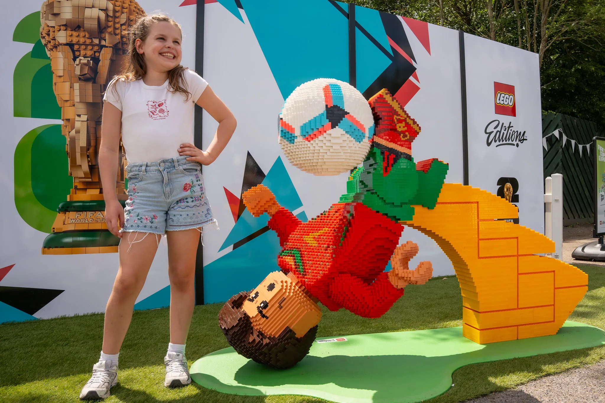 Mädchen mit weißem T-Shirt und Jeansshorts steht neben einer LEGO Figur von Christiano Ronaldo, die einen Fußball mit dem Kopf spielt, vor einer bunten Wand mit LEGO Logo und FIFA Weltmeisterschaftstrophäe aus LEGO Steinen
