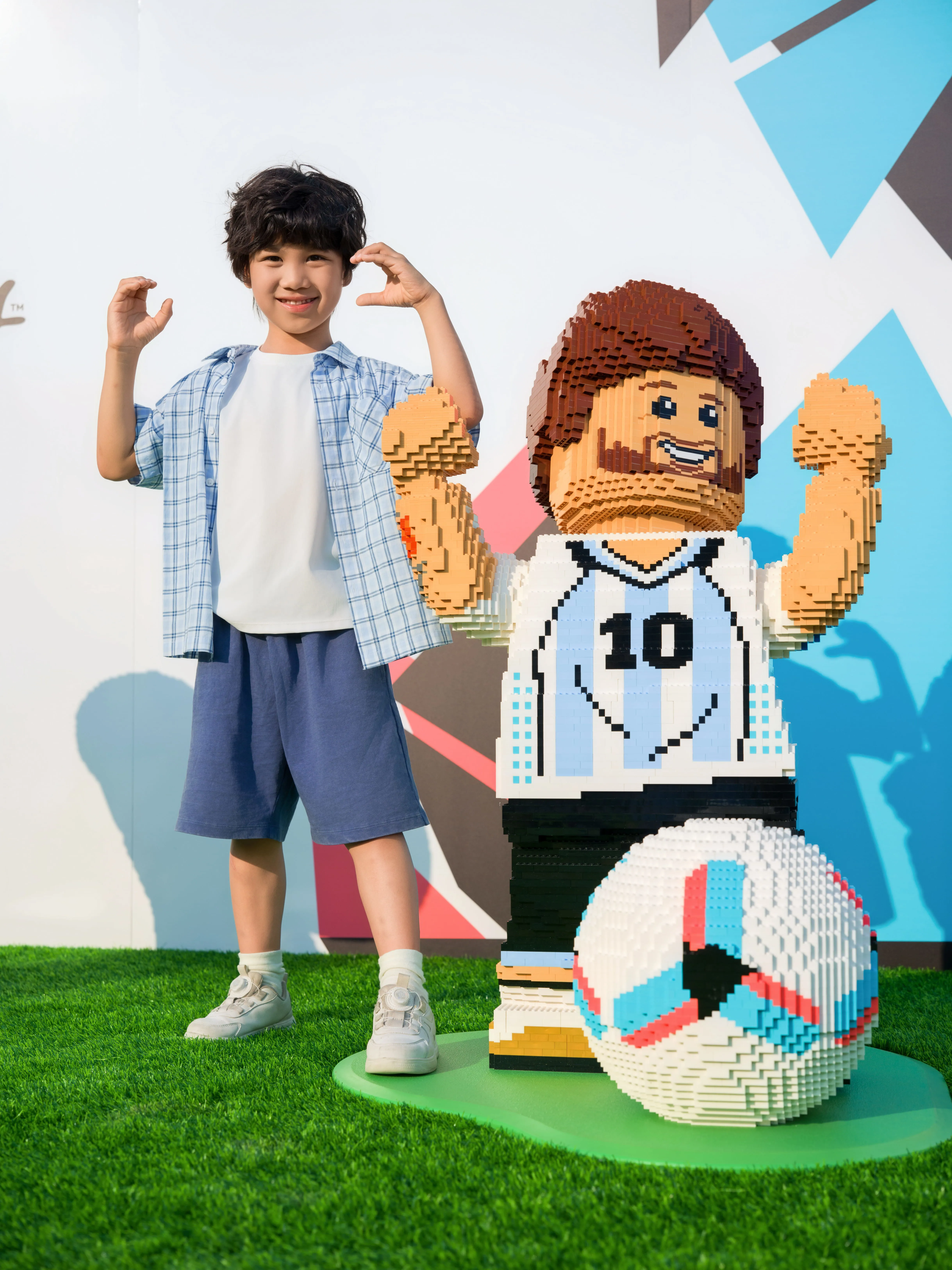 Kind steht neben einer LEGO Figur des Fußballspielers Lionel Messi mit Trikotnummer 10 und einem großen LEGO Fußball auf grünem Boden