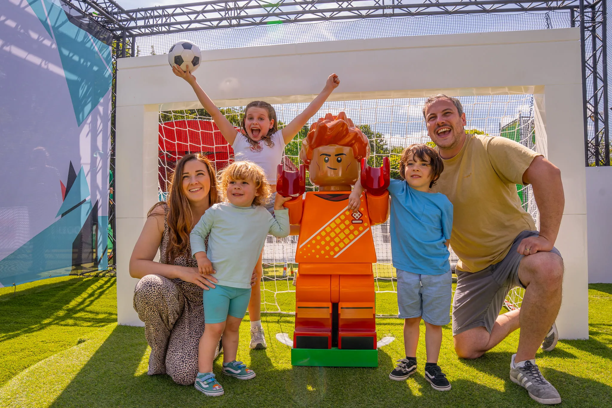 Familie mit zwei Kindern posiert neben einer großen LEGO Figur vor einem Fußballtor auf Kunstrasen