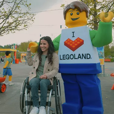 Junge Frau im Rollstuhl und ein LEGO Charakter im Eingangsbereich des LEGOLAND Deutschlands, wobei die Frau lacht, den LEGO Charakter an der Hand hält und dieser seinen Arm um ihre Schulter gelegt hat und mit dem anderen Arm nach vorne winkt.