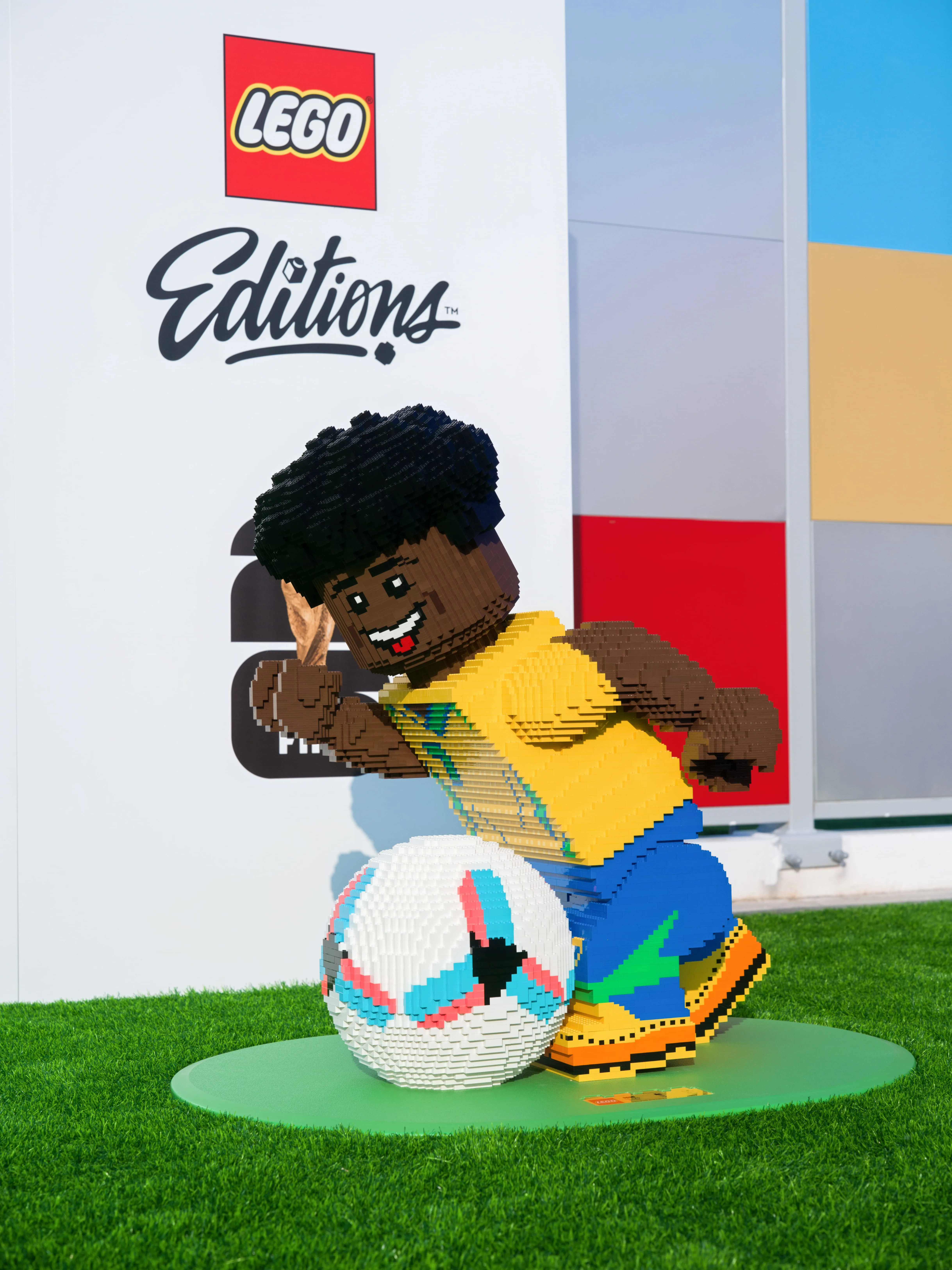 LEGO Figur aus Bausteinen mit gelbem Trikot und blau-grüner Hose, die einen Fußball auf grünem Untergrund tritt, vor einer Wand mit dem Schriftzug 'LEGO Editions'