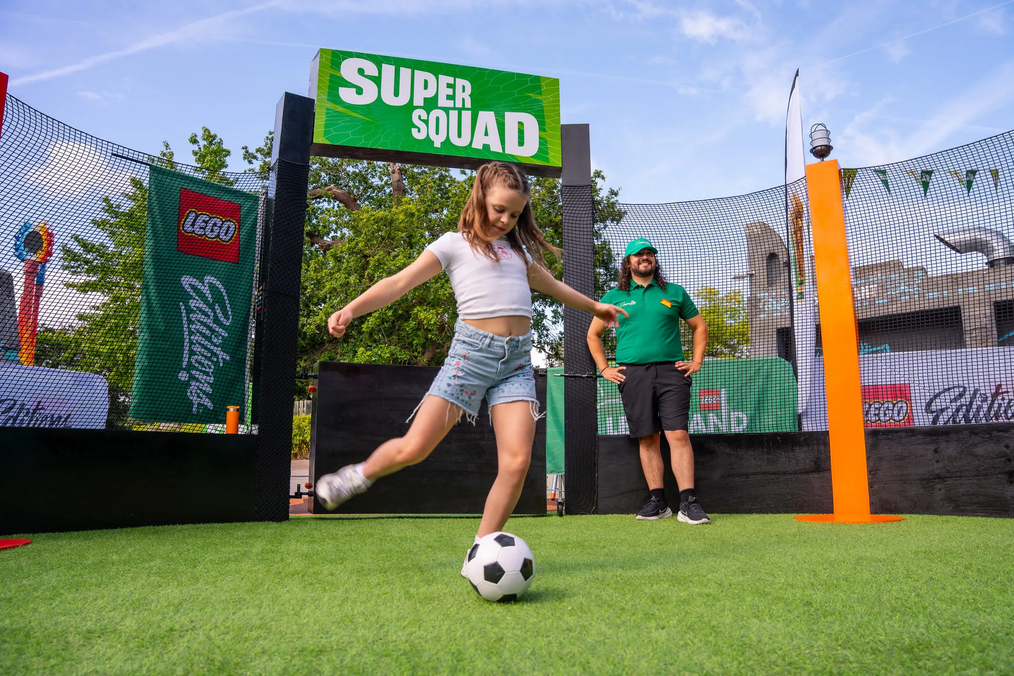 Kind in weißem T-Shirt und Jeansshorts schießt Fußball auf Kunstrasen vor grünem Schild mit Aufschrift 'SUPER SQUAD'