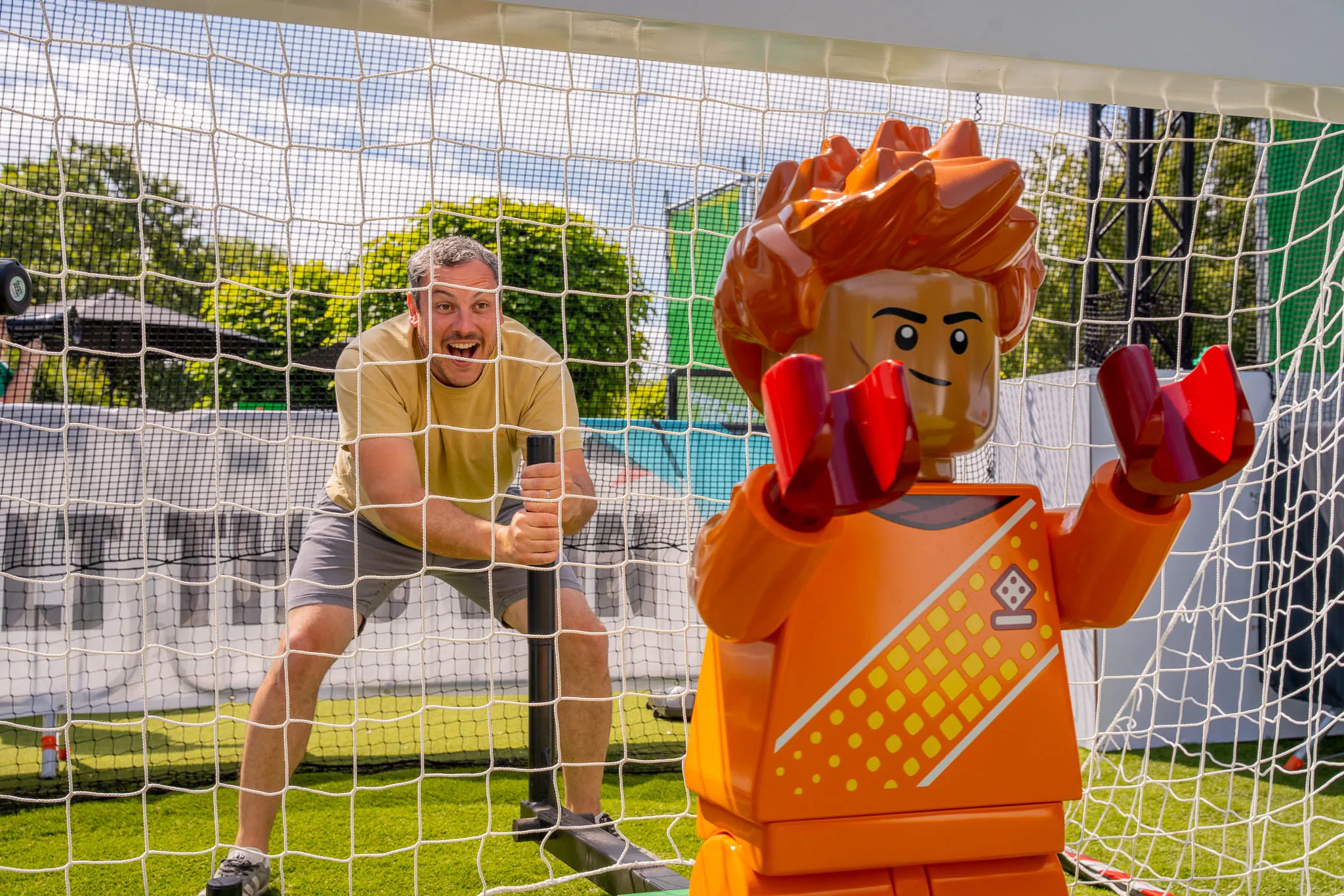 Mann in gelbem T-Shirt und Shorts steht als Torwart vor einem Fußballtor hinter einer LEGO Figur mit oranger Kleidung und roten Händen